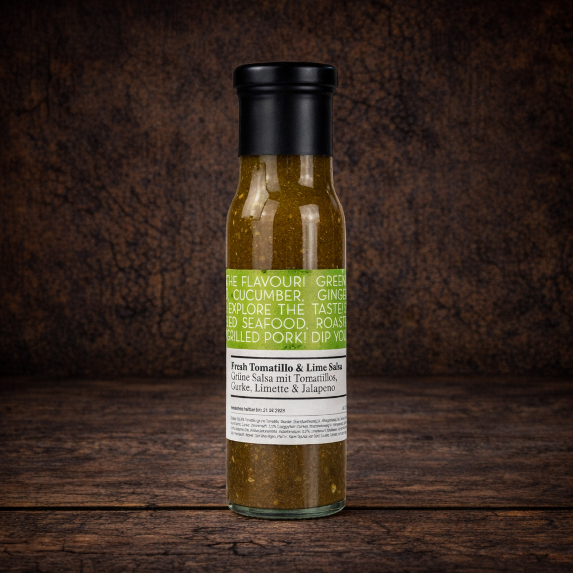 Eine schlanke Glasflasche mit schwarzem Schraubverschluss, gefüllt mit einer grünlichen, leicht körnigen Sauce. Das Etikett trägt die Bezeichnung „Fresh Tomatillo & Lime Salsa“ sowie den Hinweis „Grüne Salsa mit Tomatillos, Gurke, Limette & Jalapeño“. Die Flasche steht mittig vor einer dunklen, rustikalen Holzoberfläche.