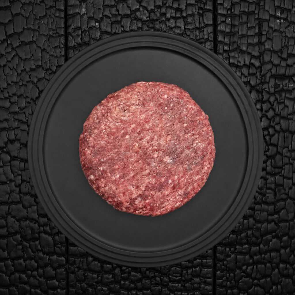 Ein rohes Wagyu Burger Patty liegt zentral auf einem runden, schwarzen Teller. Das Fleisch zeigt eine grob gewolfte Struktur mit sichtbaren hellen Fettanteilen und einer kräftig roten bis rosafarbenen Fleischfarbe. Die runde Form des Patties wirkt gleichmäßig gepresst und handwerklich hergestellt. Der Teller befindet sich auf einer schwarz verkohlten Holzoberfläche mit stark strukturierter, rissiger Maserung. Der dunkle Hintergrund hebt die Farbe und Struktur des Fleisches deutlich hervor. Die Aufnahme erfolgt aus direkter Draufsicht und wirkt reduziert, symmetrisch und grafisch. Der Fokus liegt auf der Fleischstruktur, dem Fettanteil und der hochwertigen Präsentation des Burger Patties. Die Darstellung vermittelt handwerkliche Fleischverarbeitung, hochwertige Fleischqualität und eine stilvolle Produktinszenierung.