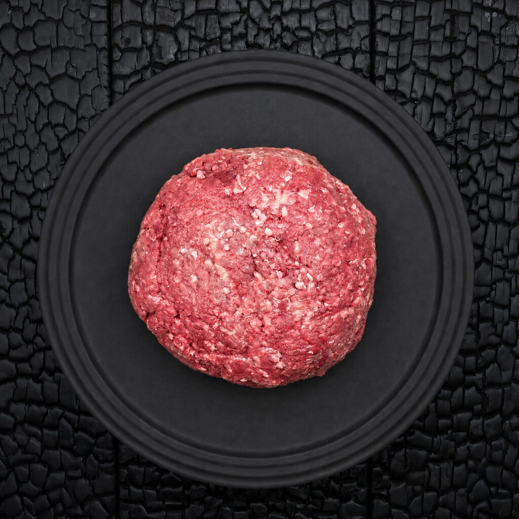 Ein großer Ballen rohes Faschiertes beziehungsweise Hackfleisch vom Wagyu Kreuzungsrind liegt zentral auf einem runden, schwarzen Teller. Das Fleisch zeigt eine grob gewolfte Struktur mit sichtbaren hellen Fettanteilen und einer kräftig roten bis rosafarbenen Farbe. Die Oberfläche wirkt locker geformt und handwerklich verarbeitet. Der schwarze Teller liegt auf einer schwarz verkohlten Holzoberfläche mit stark strukturierter, rissiger Maserung. Der dunkle Hintergrund hebt die Farbe und Struktur des Fleisches deutlich hervor und erzeugt einen starken Kontrast. Die Aufnahme erfolgt aus direkter Draufsicht und wirkt reduziert, symmetrisch und grafisch. Der Fokus liegt auf der Fleischstruktur, dem Fettanteil und der hochwertigen Präsentation des frischen Hackfleischs. Die Darstellung vermittelt handwerkliche Fleischverarbeitung, hochwertige Fleischqualität und eine stilvolle Produktinszenierung.